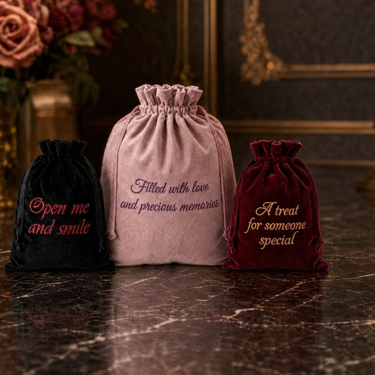 Personalised luxury velvet gift bags set in black, dusty pink, and burgundy with custom flat-stitch embroidery reading 'Open me and smile', 'Filled with love and precious memories', and 'A treat for someone special', centered on a dark marble surface.